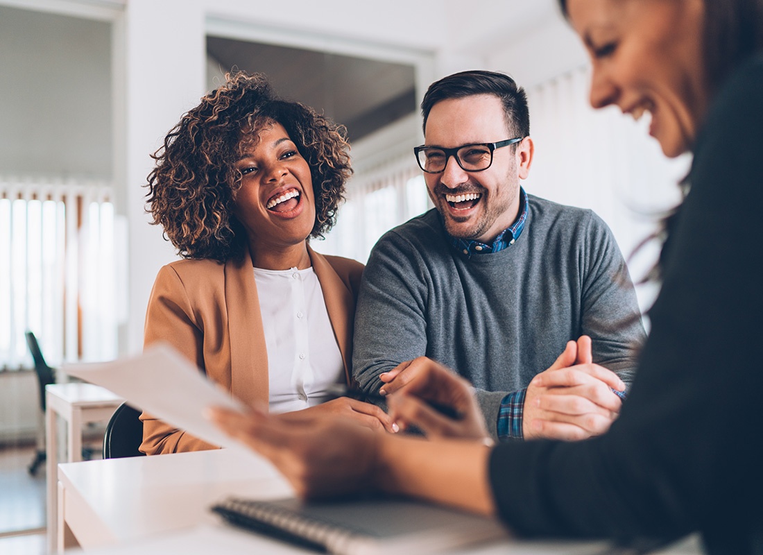 About Our Agency - Portrait of a Cheerful Diverse Young Couple Meeting with a Female Agent Holding Documents in a Small Office